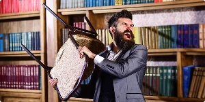 A bearded man in a suit stands angrily holding a chair, as if preparing to throw it.