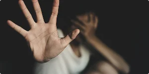 A person with a pained expression raises one hand to block the camera while hiding their face with the other hand.