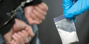 A person in blue gloves holds a bag of white powder, with handcuffed hands visible in the background.