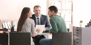 A man and woman sit at a table with a suited man, engaged in conversation in a professional setting.