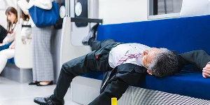 A man in a suit and tie lies on a blue metro bench, appearing unwell or possibly intoxicated.