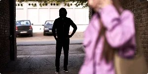 A woman walks through a dimly lit alley, unaware of a hooded man stalking her from behind in the narrow space.