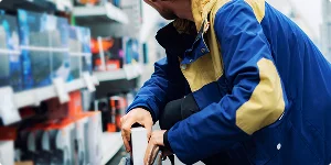 A man in a blue jacket appears to be stealing an item from a store shelf while looking around cautiously.