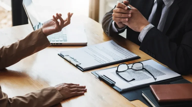 Two people chatting at a wooden desk with papers, glasses, and a laptop nearby.
