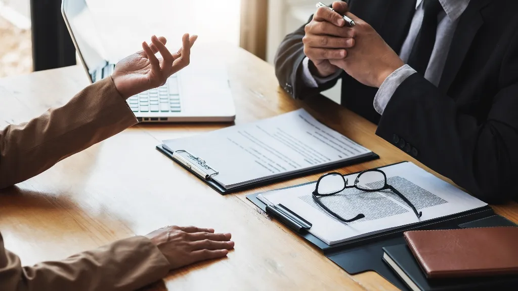 Two people chatting at a wooden desk with papers, glasses, and a laptop nearby.
