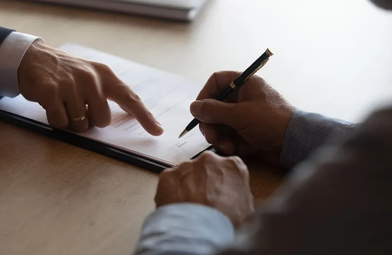 Two people at a table, one pointing at a paper, the other holding a pen ready to sign.