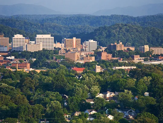 A sunny cityscape with lots of green trees and mountains in the background.
