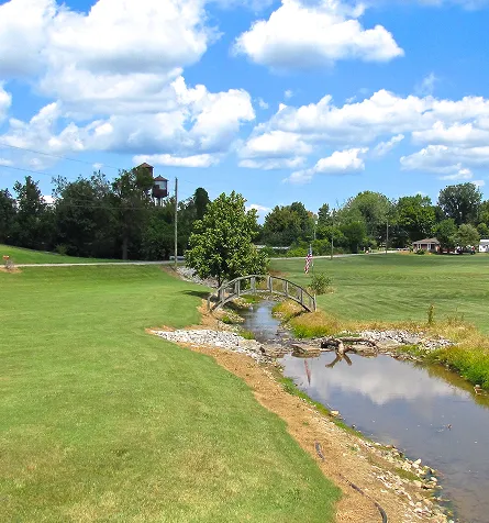 A sunny park with a small wooden bridge over a creek and green grass everywhere.