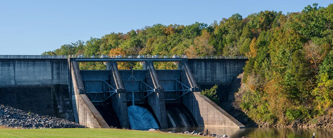 A big concrete dam with water flowing through one gate, surrounded by green and autumn trees.