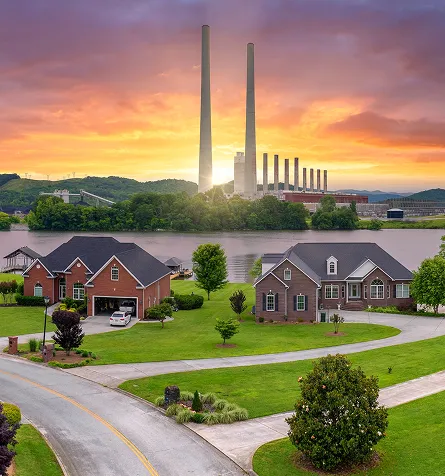 Cozy suburban homes with green lawns by a river, industrial smokestacks glowing in sunset.