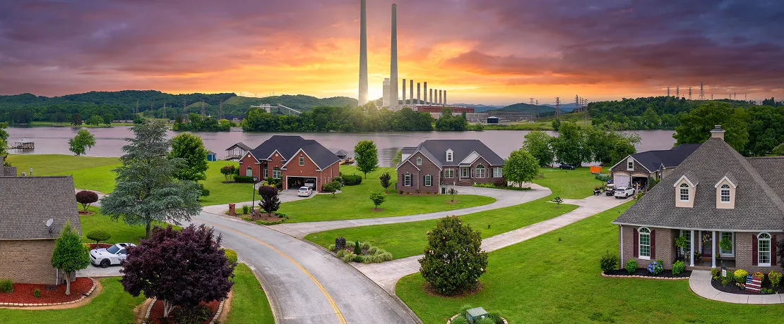 Cozy neighborhood with green lawns by a river, colorful sunset, and tall factory chimneys.