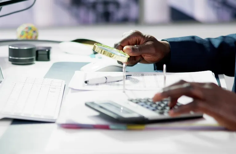 A person using a magnifying glass and calculator at a white desk.
