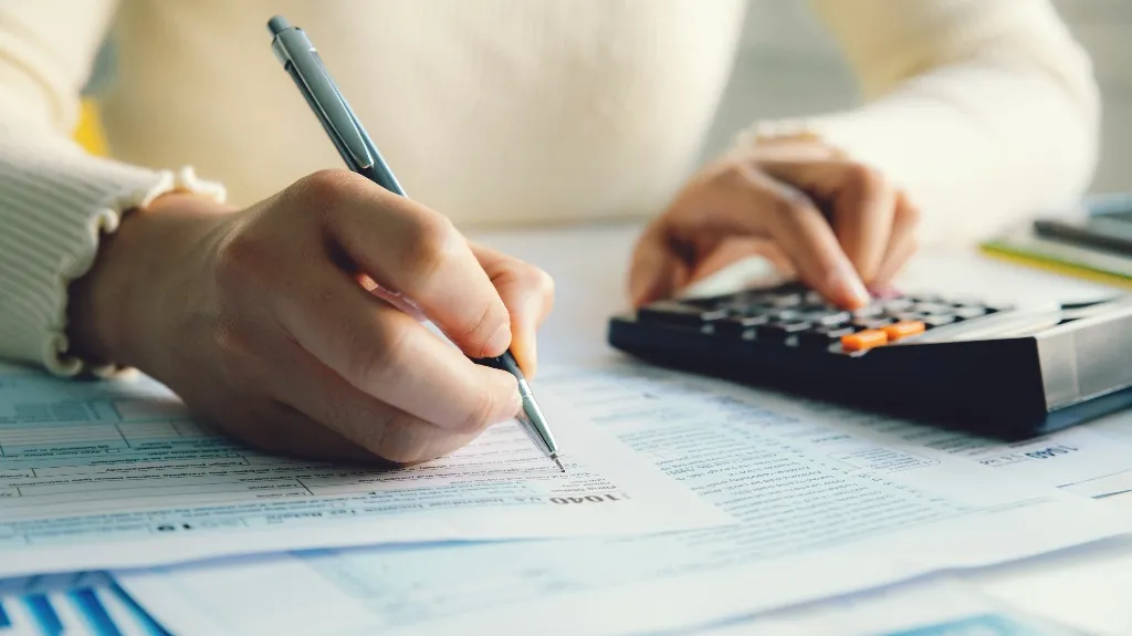 A person’s hands filling out tax forms with a pen and using a black calculator.