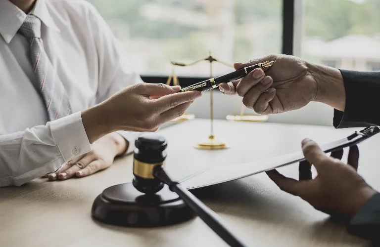 Two people exchanging a black pen over a desk with a gavel and scales in the background.