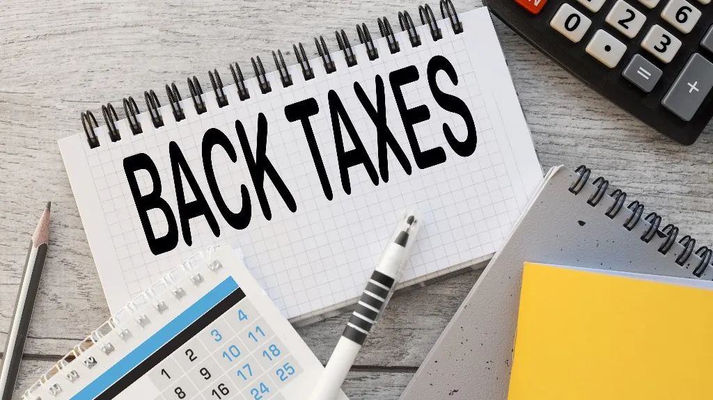 A notebook with "BACK TAXES" written, surrounded by a pen, calendar, pencil, and calculator.