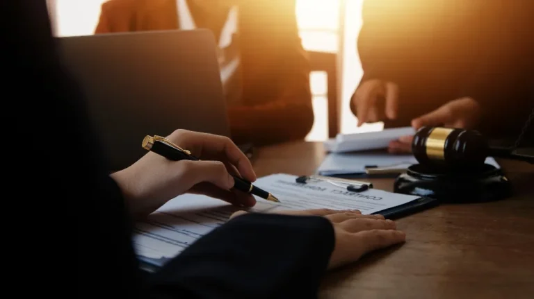 A person signing a contract with a pen, a gavel and laptop nearby.