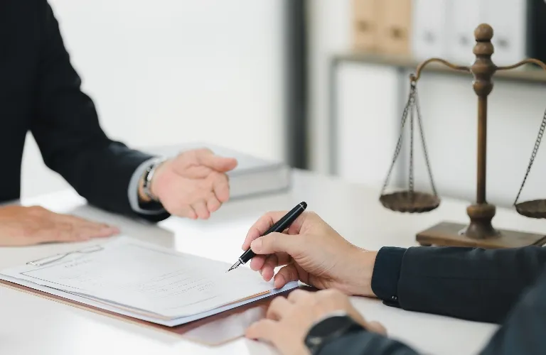 Two people in black suits chatting with a pen and papers on a white desk, and a bronze scale nearby.