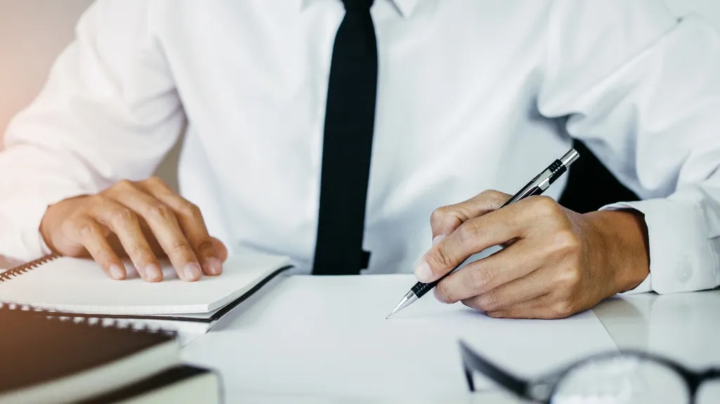 Person in white shirt and black tie writing on paper with pen, notebook nearby.