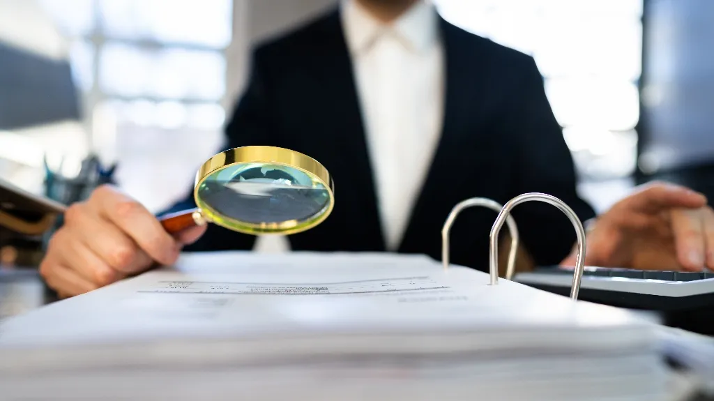 Person in black suit checking papers with a gold-rimmed magnifying glass and calculator.