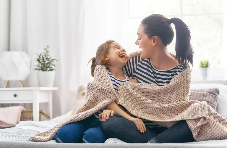 A mom and daughter in striped shirts laughing, wrapped in a cozy beige blanket.