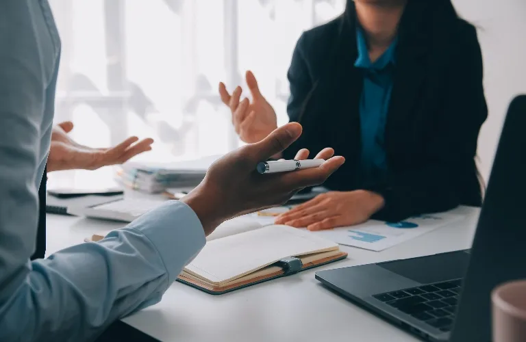 Two people chatting at a desk with a notebook, laptop, and papers around.
