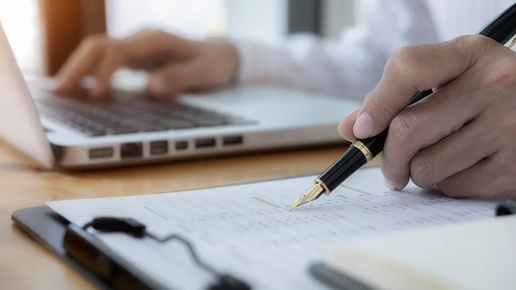 Close-up of a hand writing with a fancy black pen near a laptop on a wooden table.