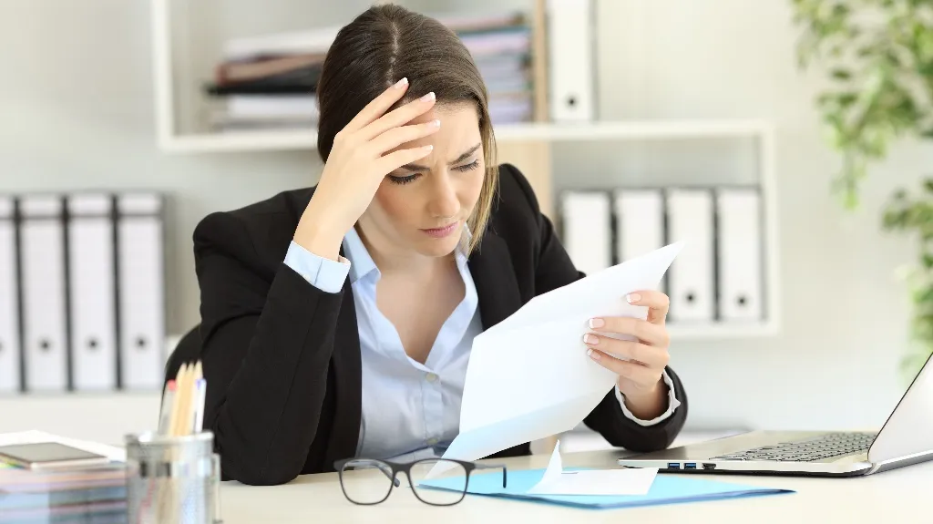 A woman in a black blazer looks stressed while reading a paper at her desk.