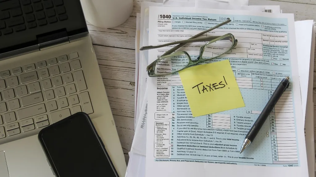 A laptop, smartphone, glasses, pen, and tax forms with a sticky note saying "TAXES!