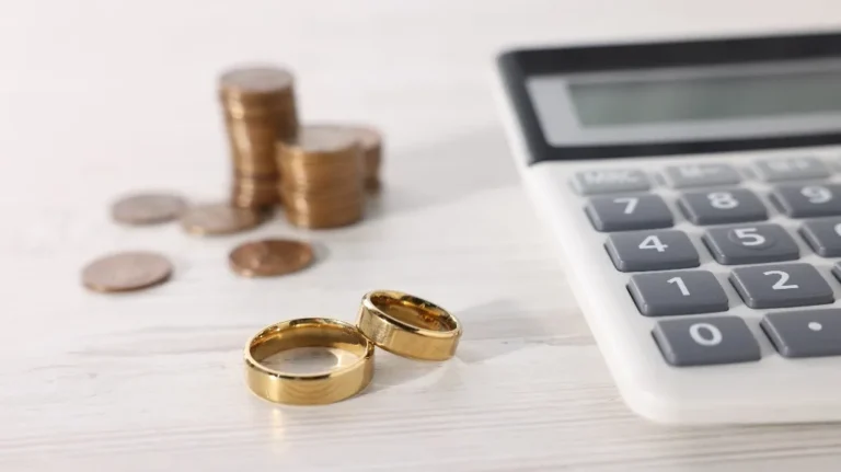 Two shiny gold rings, a white calculator, and stacks of coins on a wooden table.