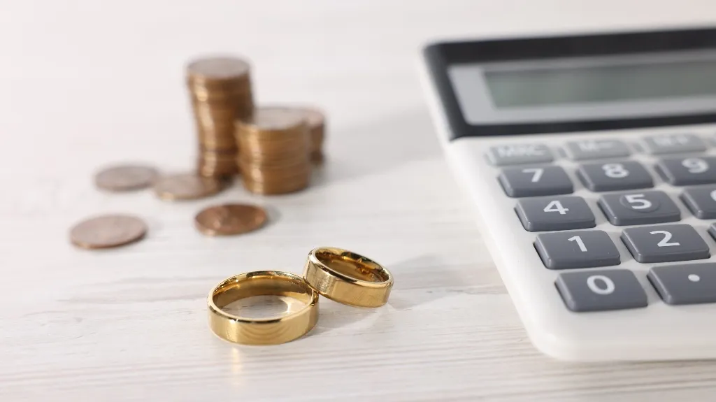 Two shiny gold rings, a white calculator, and stacks of coins on a wooden table.