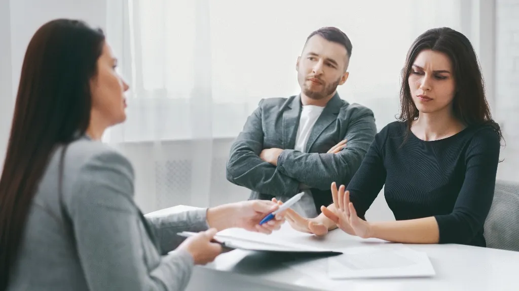 Three people having a serious discussion at a white table with papers and a pen.