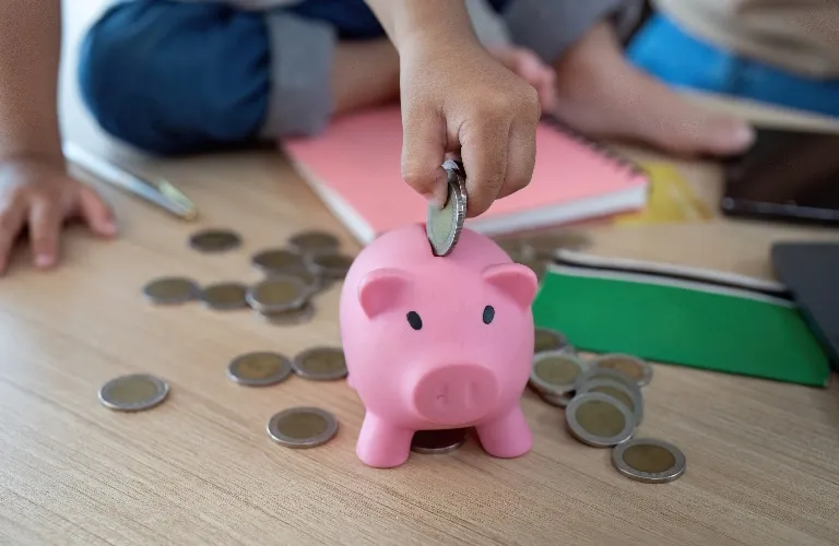 A kid's hand dropping a coin into a cute pink piggy bank surrounded by scattered coins.