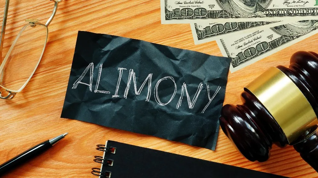 A crumpled black paper with "ALIMONY" written on it, money, judge's gavel, glasses, and pen on wooden table.