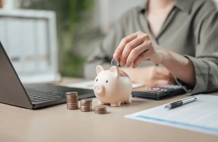Someone dropping a coin into a cute pink piggy bank by a laptop and papers.
