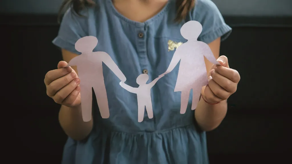 Kid in blue dress holding paper cutout of a family with two adults and a child.