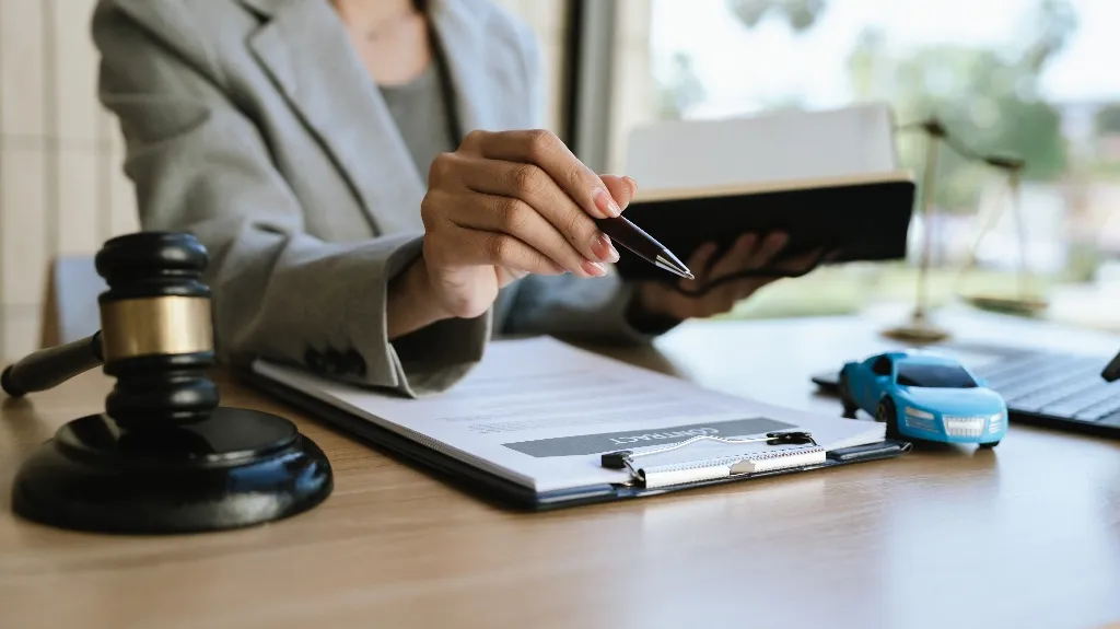 Person in gray blazer holding pen and book, with gavel, contract, and toy blue car on desk.