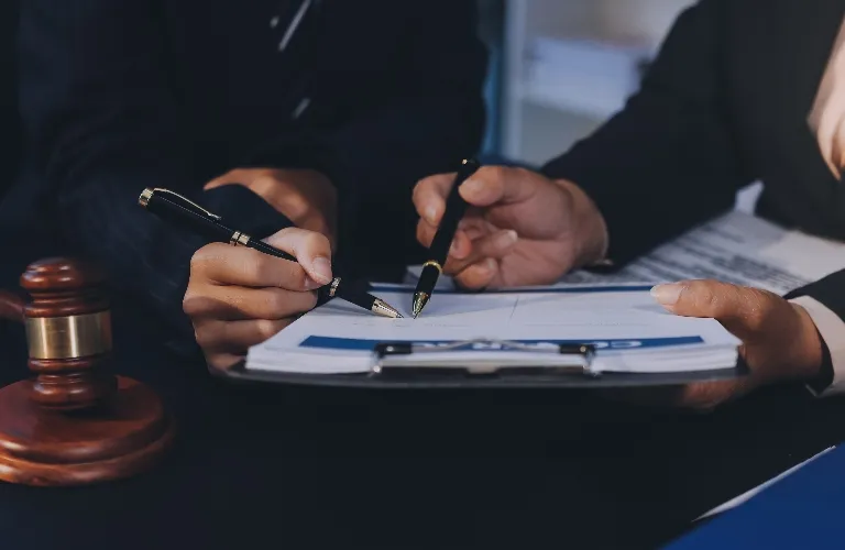 Two people in suits pointing at papers on a clipboard with pens, wooden gavel nearby.
