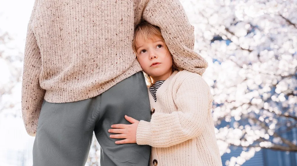 Little kid in cream sweater hugging adult's leg with soft white blossoms in background.