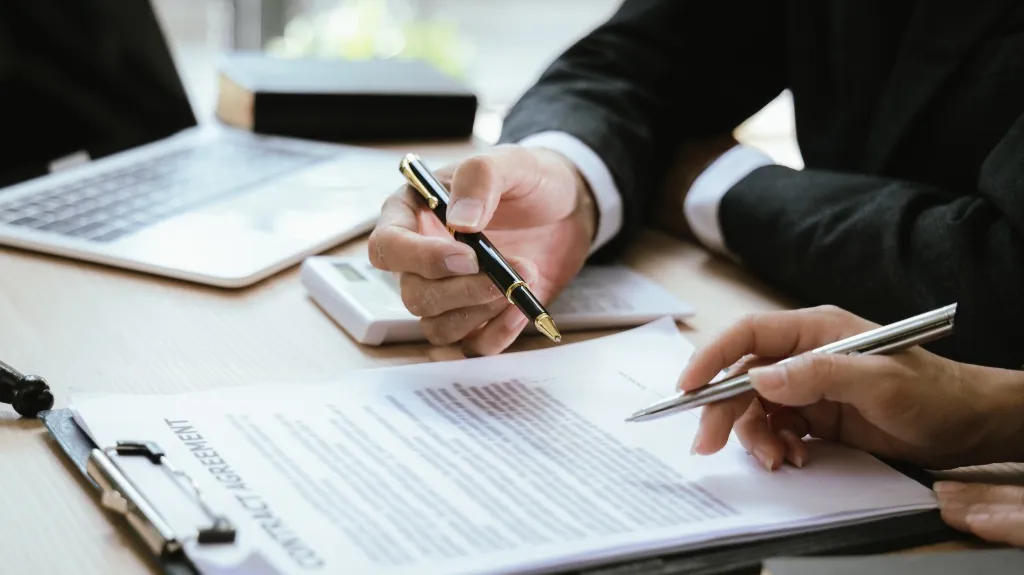 Two people in suits pointing at a contract agreement with pens on a desk.