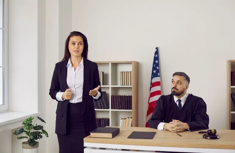 A woman lawyer talking while a judge listens at a desk with an American flag.