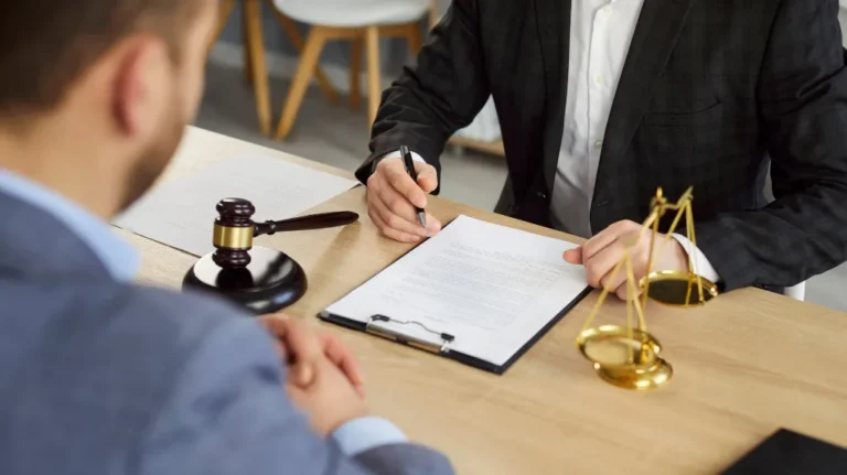 Two people chatting at a desk with legal papers, a gavel, and golden scales.