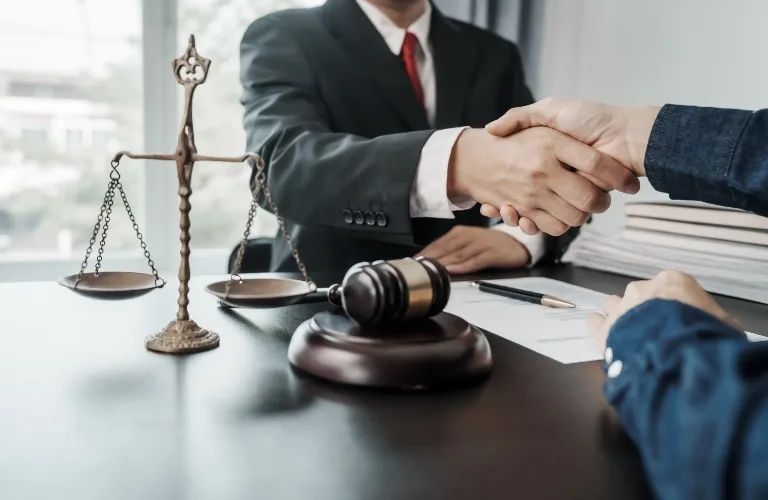 Two people shaking hands over a desk with a gavel and scales of justice nearby.