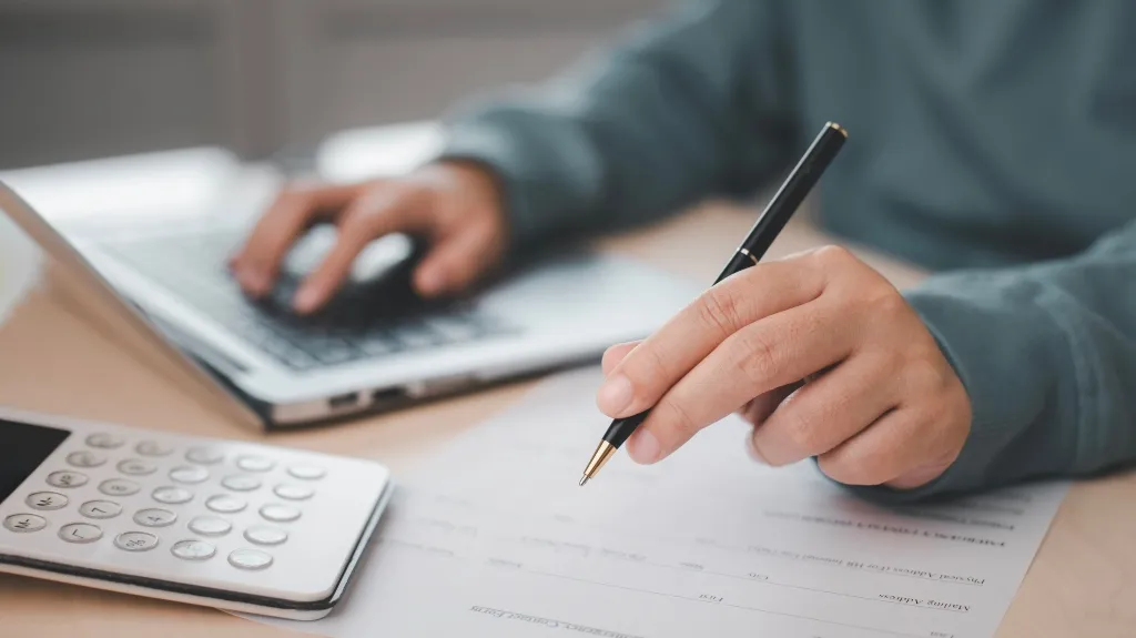 Person in a blue sweatshirt working with a laptop, pen, paper, and calculator.