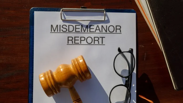Clipboard with "MISDEMEANOR REPORT" paper, wooden gavel, and black glasses on a wooden table.