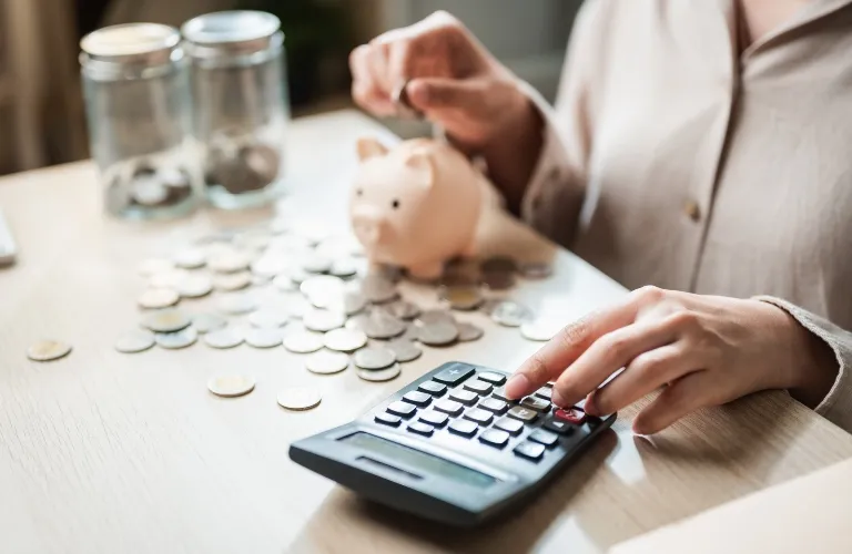Person using a calculator with coins, piggy bank, and jars on a wooden table.