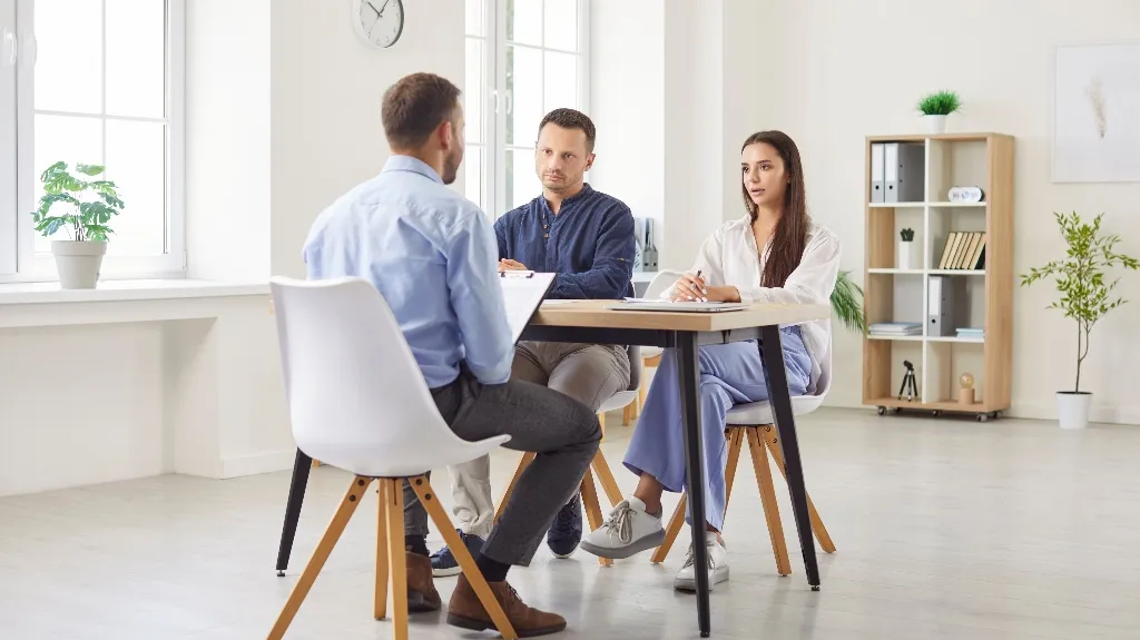 Three people chatting in a bright office with plants and a clock on the wall.