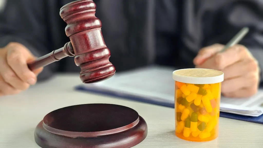 A judge’s hand holding a wooden gavel near a pill bottle filled with yellow and green capsules.