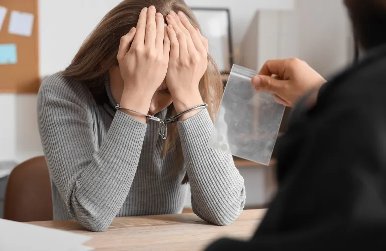 A woman in handcuffs covering her face while someone shows a small plastic bag.