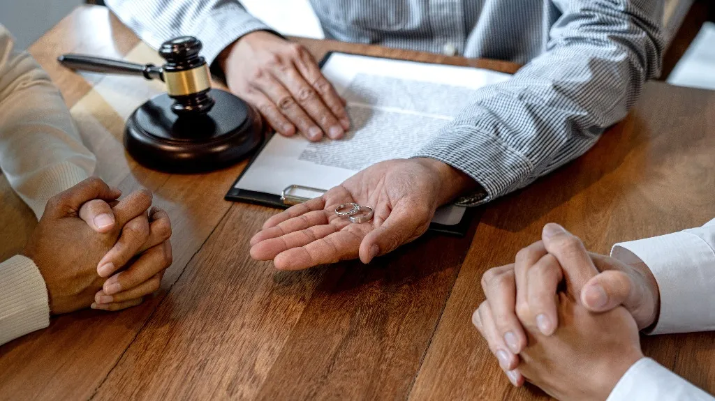 Three people at a wooden table with wedding rings, a gavel, and legal papers.