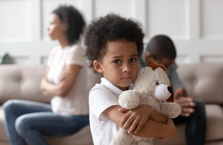 A sad little boy hugging a teddy bear with two adults sitting upset in the background.